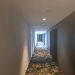 Długi korytarz hotelowy z wykładziną w geometryczne wzory, oświetlony punktowymi lampami sufitowymi i światłem z końca korytarza.