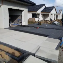 Układanie jasnoszarej kostki brukowej na podjeździe do garażu, z widoczną maszyną do ubijania i robotnikiem w tle, na tle budynków w stanie surowym.