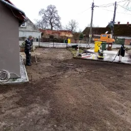 Przygotowanie terenu pod przyszły podjazd: wyrównana ziemia, widoczny pracownik z łopatą, częściowo ułożony obrzeżnik, narzędzia i materiały budowlane w tle, w tym zagęszczarka i taczka z piaskiem.