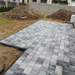 Nowo ułożony taras z szarej kostki brukowej na piasku, widoczne hałdy ziemi w tle i fragment budynku z klimatyzatorem.