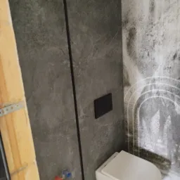 Wnętrze łazienki w trakcie wykańczania: widoczna podtynkowa spłuczka WC z czarnym przyciskiem, fragment ściany z szarą płytką imitującą kamień i fragment ściany z wzorem imitującym zacieki, a także...