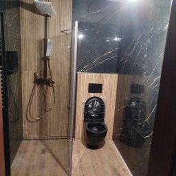 USŁUGI REMONTOWO WYKOŃCZENIOWE MARCIN LEWANDOWSKI - Nowoczesna łazienka z czarną armaturą i panelami drewnopodobnymi, widoczna kabina prysznicowa i podwieszana toaleta w folii ochronnej.