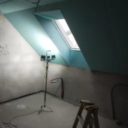 Poddasze w trakcie adaptacji: widoczne płyty kartonowo-gipsowe na skosach, okno dachowe, lampa budowlana na statywie, drabina z narzędziami, surowe ściany i podłoga.