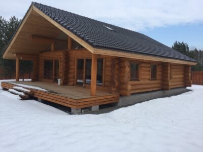 Gotowy parterowy dom z bali z ciemną dachówką, drewnianym tarasem i schodami, otoczony śniegiem.