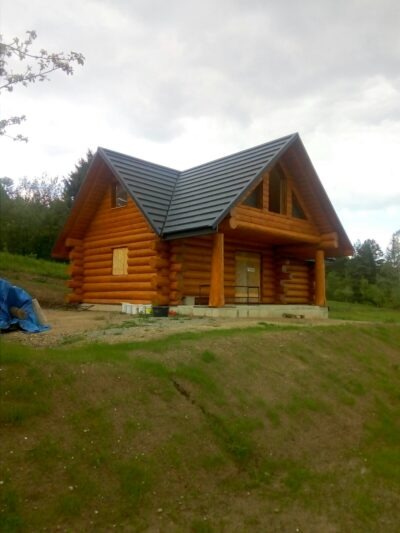 Nowo wybudowany, parterowy dom z bali, z ciemnoszarą, metalową dachówką, widoczny z dołu, na tle zieleni i pochmurnego nieba. Na pierwszym planie niewielkie nierówności terenu.