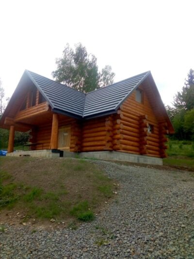 Gotowy, parterowy dom z bali z ciemnoszarą, metalową dachówką, położony na zboczu z kamienistym podjazdem.