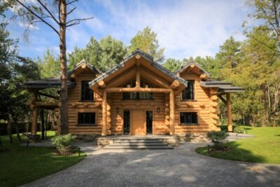 Dwukondygnacyjny dom z bali z ciemnymi oknami i szarym brukowanym podjazdem, otoczony drzewami i zielenią.