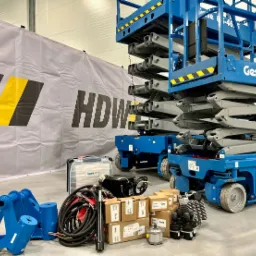 Niebieski podest ruchomy Genie GS-1932, części zamienne i skrzynka narzędziowa na tle szarego banera HDW w hali.