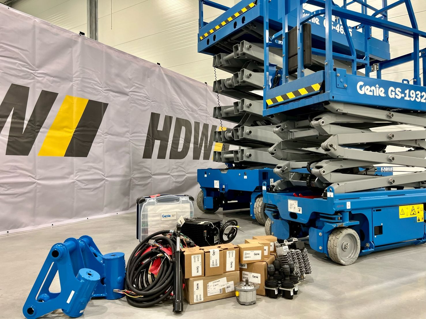 Niebieski podest ruchomy Genie GS-1932, części zamienne i skrzynka narzędziowa na tle szarego banera HDW w hali.