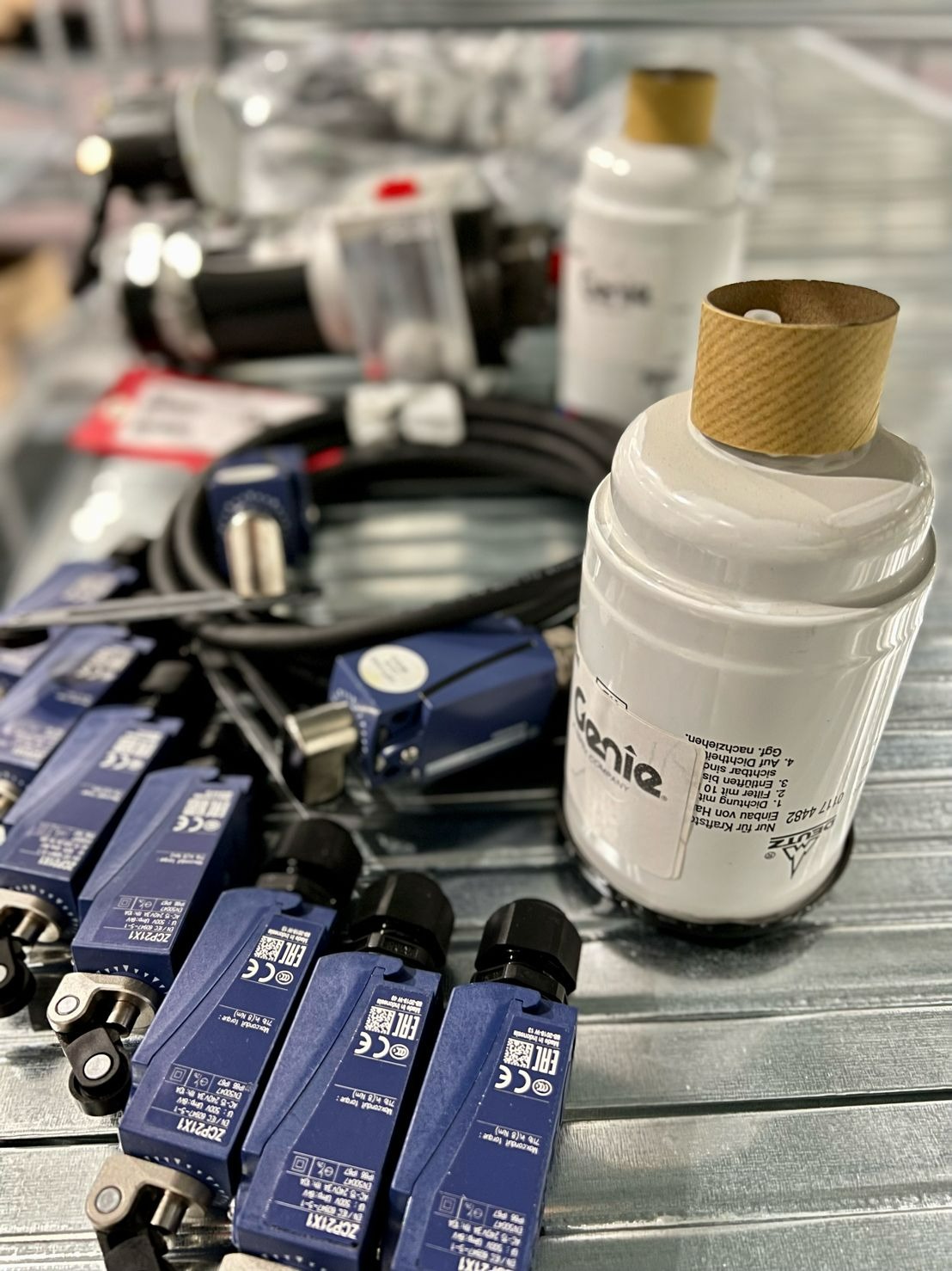 Zbliżenie na nowe czujniki krańcowe, przewody elektryczne i filtry hydrauliczne marki Genie na metalowej powierzchni, prawdopodobnie przygotowane do montażu lub serwisu.