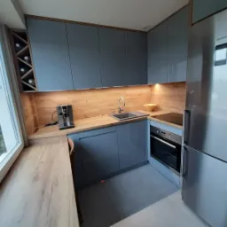 Nowoczesna kuchnia w odcieniach szarości z drewnianymi akcentami, oświetleniem LED pod szafkami i widokiem na miasto przez okno.