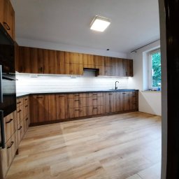 Meblotoss Jacek Owca - Nowoczesna kuchnia z drewnianymi frontami szafek, czarnym blatem i białymi płytkami na ścianie, widziana zza framugi drzwi.