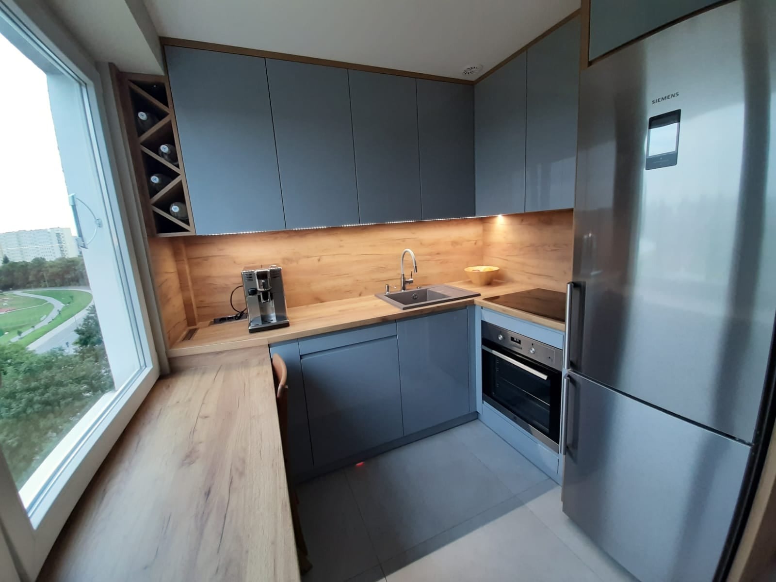 Nowoczesna kuchnia w odcieniach szarości z drewnianymi akcentami, oświetleniem LED pod szafkami i widokiem na miasto przez okno.