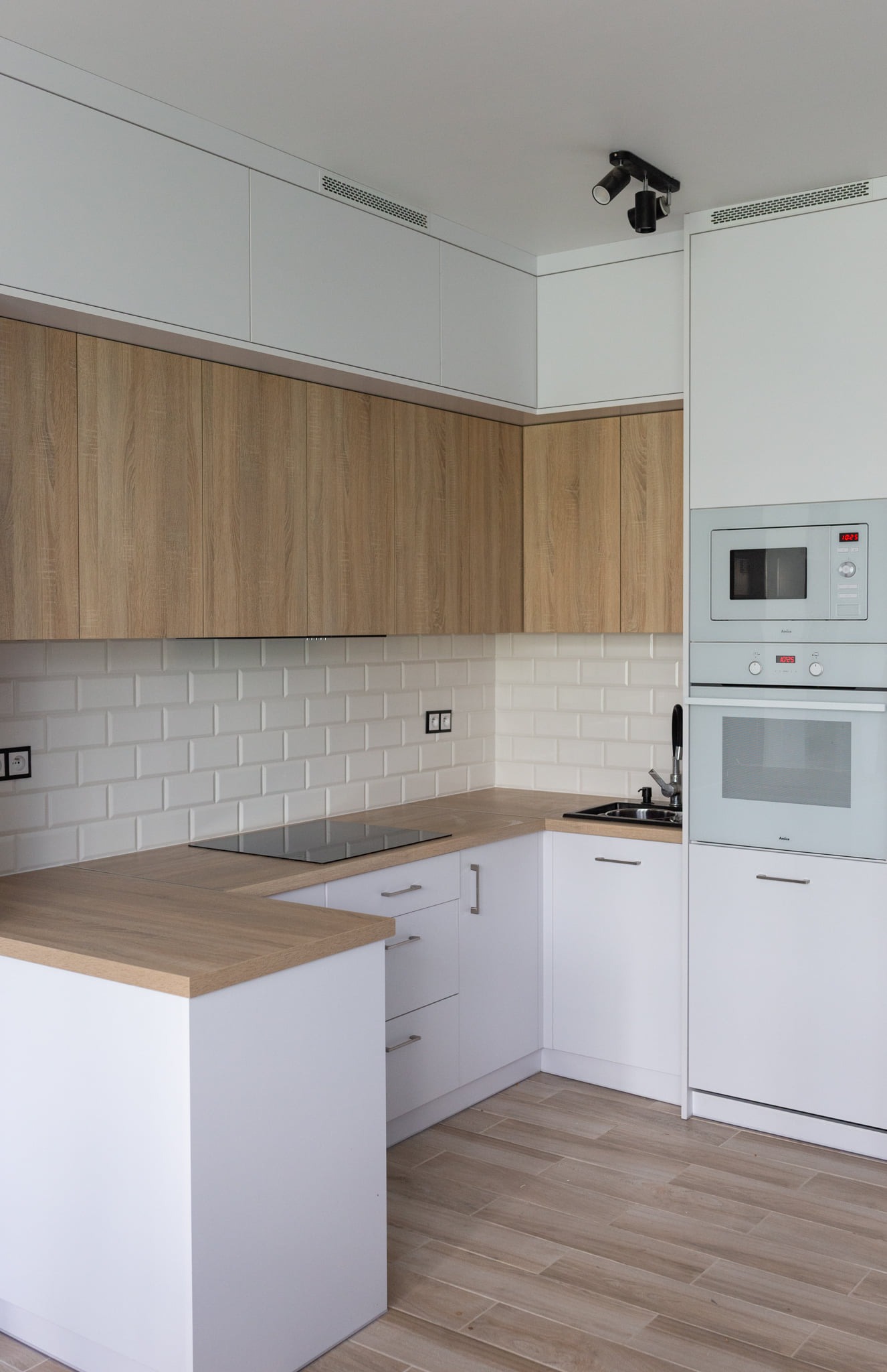Jasna kuchnia w stylu skandynawskim z drewnianymi blatami i frontami szafek górnych, białe płytki na ścianie, minimalistyczne oświetlenie podszafkowe.