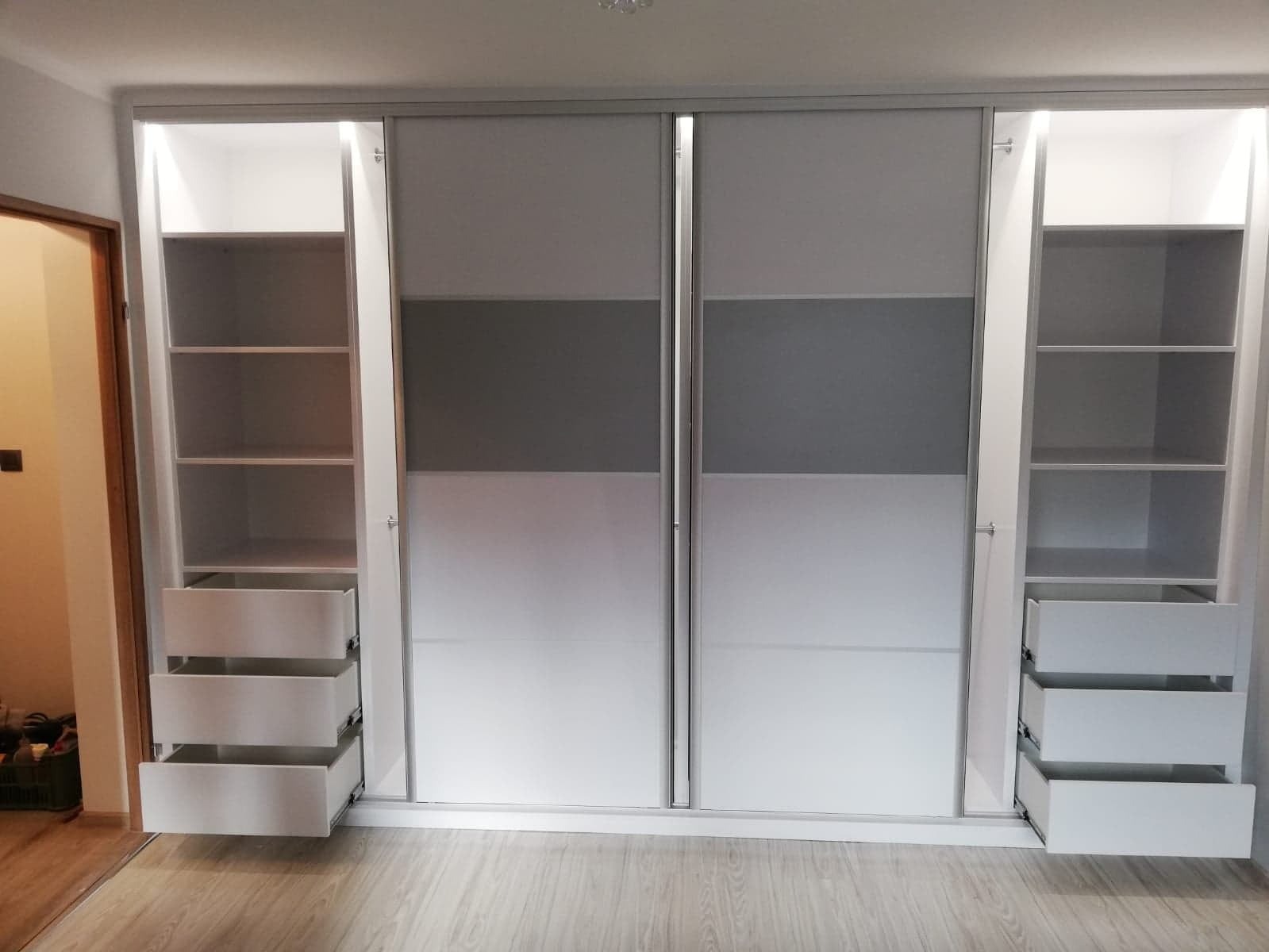 Biała szafa przesuwna z szarymi elementami, oświetleniem wewnętrznym i wysuwanymi szufladami, widoczna z przodu, z otwartymi drzwiami po bokach.