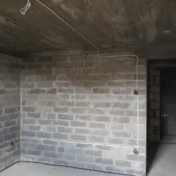 Surowy mur z bloczków betonowych z widocznymi przewodami elektrycznymi i puszkami instalacyjnymi w trakcie montażu, widok wnętrza w stanie deweloperskim.