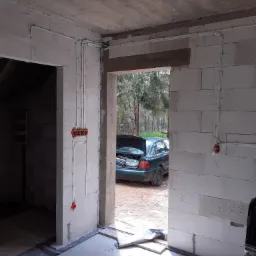 Wnętrze surowego budynku z widoczną instalacją elektryczną w peszlach i puszkach, samochód z otwartym bagażnikiem w tle.