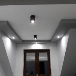 Nowoczesne oświetlenie sufitowe z trzema punktami świetlnymi w czarnych oprawach, zamontowane na podwieszanym suficie z szarymi elementami dekoracyjnymi, nad drewnianymi drzwiami balkonowymi...