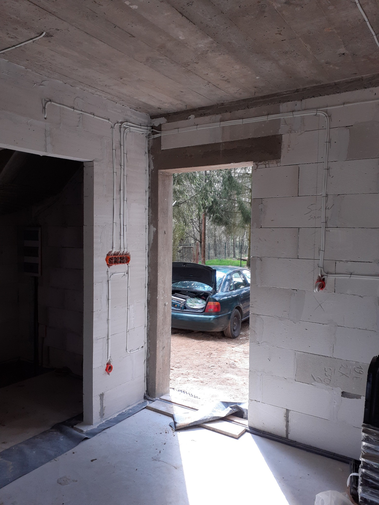 Wnętrze surowego budynku z widoczną instalacją elektryczną w peszlach i puszkach, samochód z otwartym bagażnikiem w tle.
