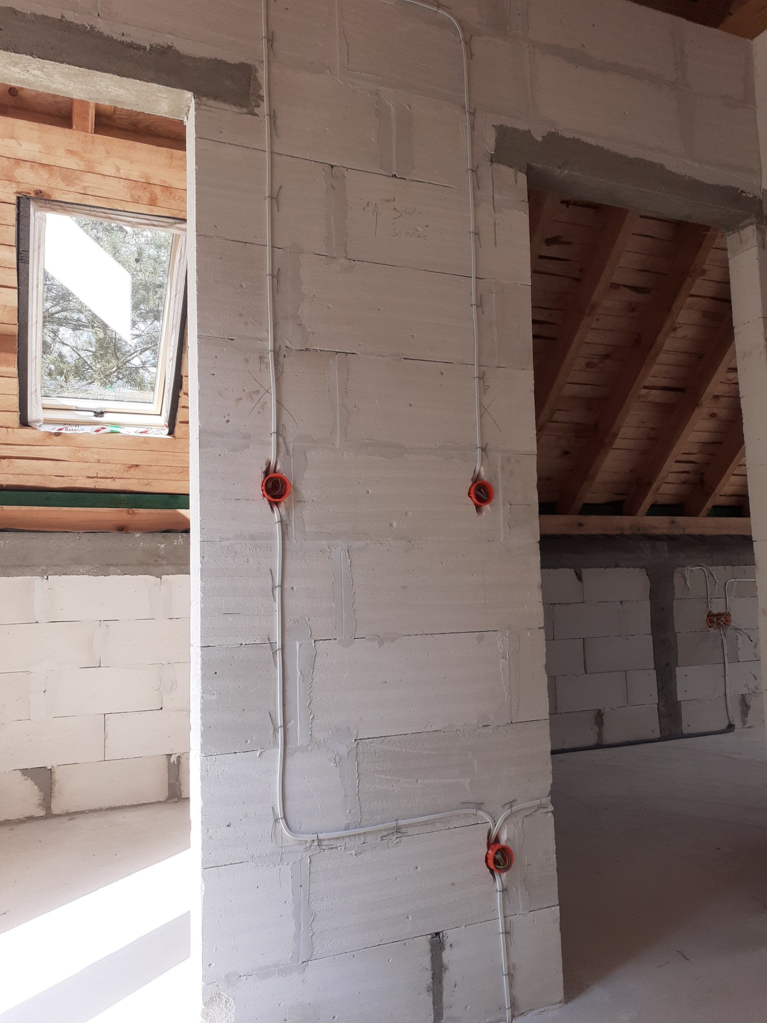 Wewnętrzna instalacja elektryczna w trakcie montażu w surowym budynku z białych bloczków, widoczne kable i pomarańczowe puszki instalacyjne, drewniana konstrukcja dachu i okno dachowe w tle.