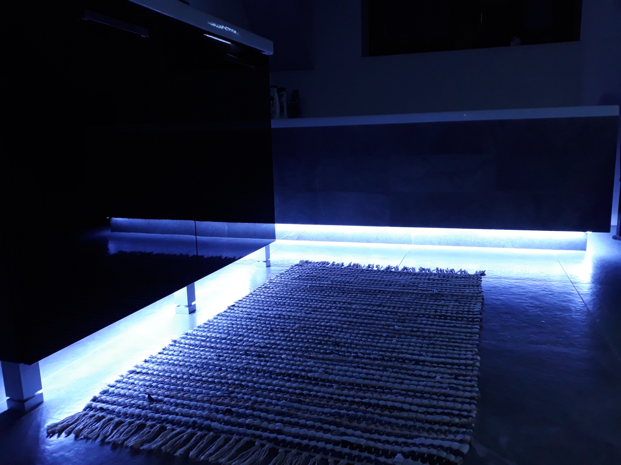 Nowoczesna kuchnia z podświetleniem LED pod szafkami, oświetlająca dywanik z frędzlami na błyszczącej podłodze.