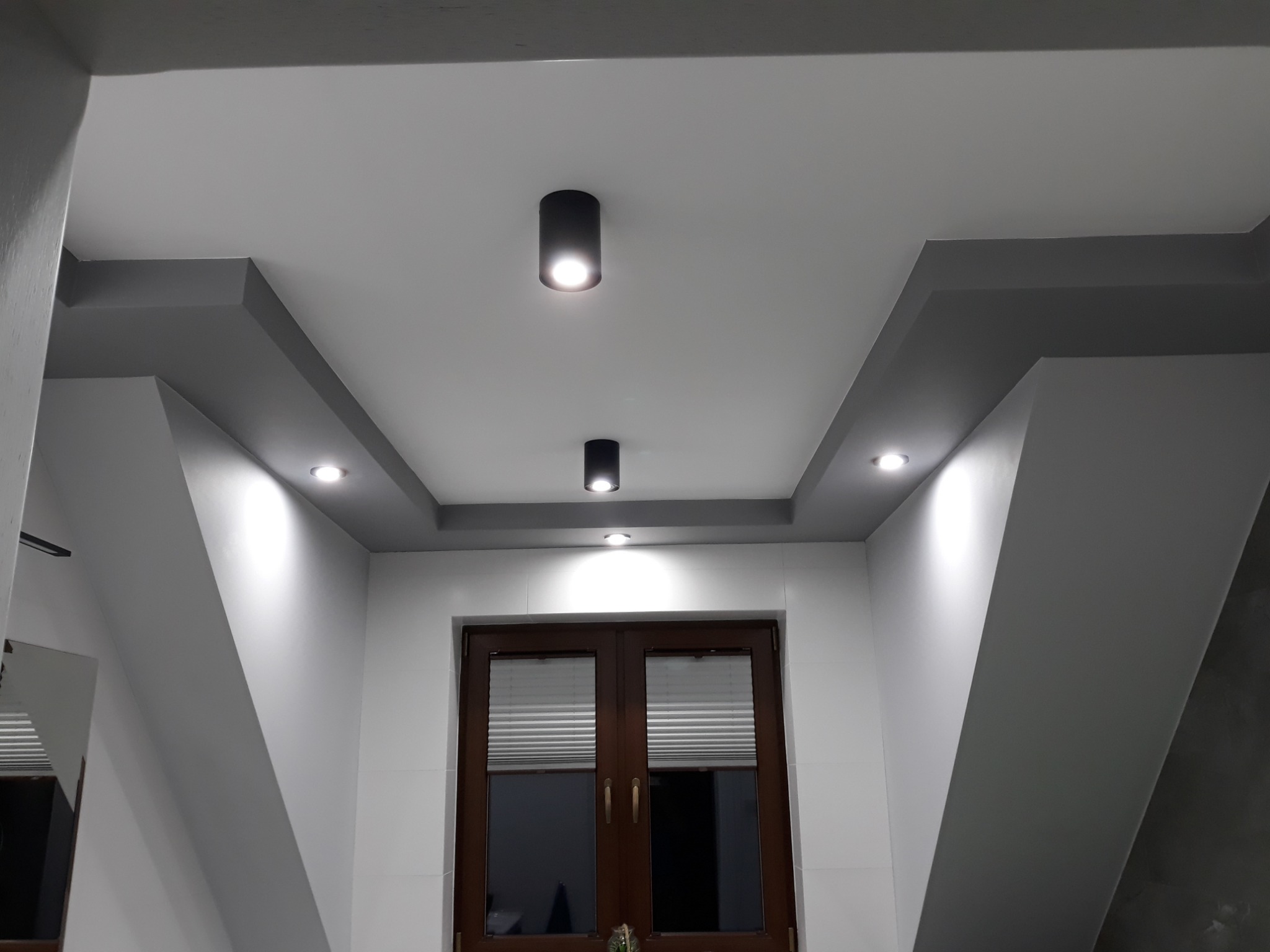 Nowoczesne oświetlenie sufitowe z trzema punktami świetlnymi w czarnych oprawach, zamontowane na podwieszanym suficie z szarymi elementami dekoracyjnymi, nad drewnianymi drzwiami balkonowymi...