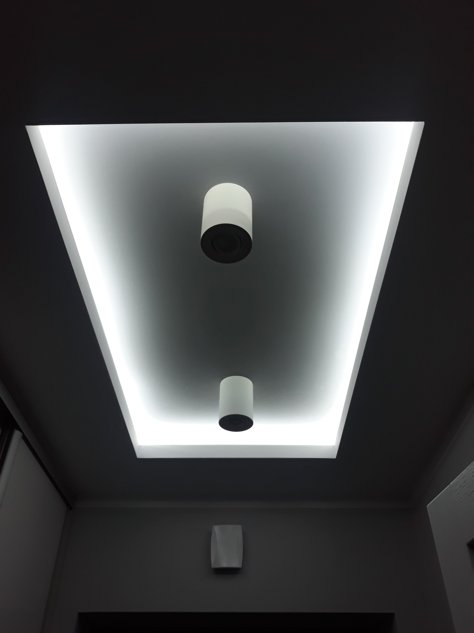 Nowoczesny sufit podwieszany z oświetleniem LED w kształcie trapezu i dwoma cylindrycznymi lampami wiszącymi na tle szarej ściany z widocznym elementem wentylacyjnym.