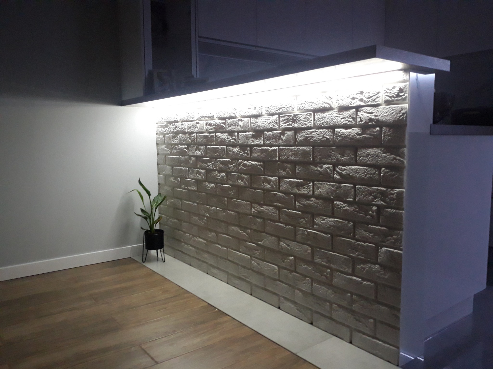 Podświetlona ściana z cegły dekoracyjnej w salonie, z małą rośliną w czarnej doniczce i drewnianą podłogą.