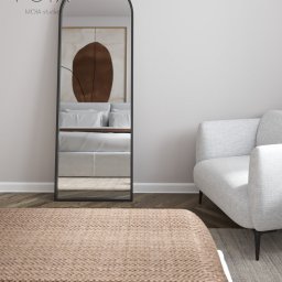 MOIA studio - Sypialnia z dużym lustrem w czarnej ramie, odbijającym łóżko z obrazem na ścianie, jasnoszary fotel na tle jasnej ściany i drewnianej podłogi.