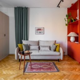 Fresh Home - Jasny salon z parkietem w jodełkę, szarą sofą z poduszkami, stolikiem z rogalikiem, zielonymi zasłonami i pomarańczową zabudową ścienną z półkami.