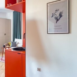 Fresh Home - Częściowy widok pomarańczowej zabudowy kuchennej z czarnym zlewem w nowoczesnym wnętrzu z białymi ścianami i podłogą w terazzo; w tle widoczny salon z turkusowymi zasłonami.