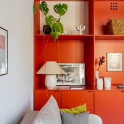 Fresh Home - Pomarańczowa zabudowa meblowa w salonie z dekoracjami: lampka nocna, roślina Monstera, koszyk wiklinowy i grafiki na ścianie.