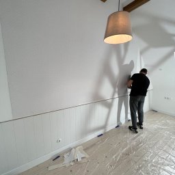 Fresh Home - Pracownik wygładza tapetę na ścianie w pokoju z drewnianą podłogą zabezpieczoną folią malarską, widoczne listwy przypodłogowe i lampa wisząca.