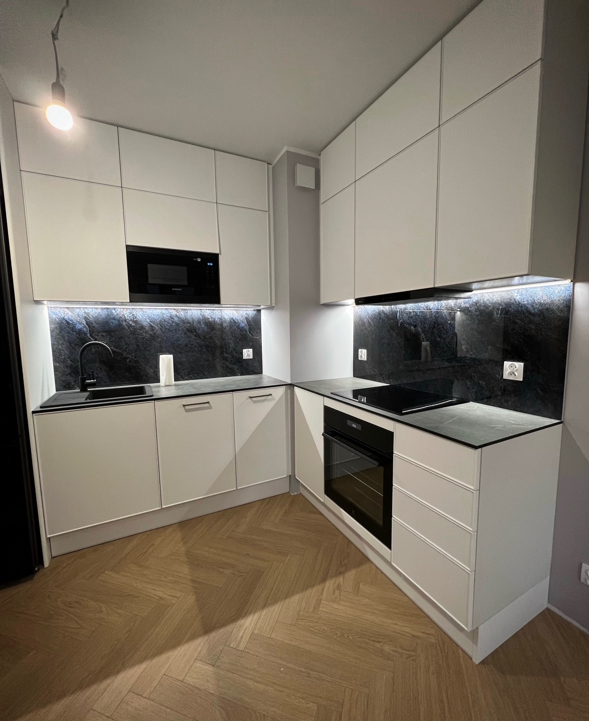 Nowoczesna kuchnia z białymi szafkami, czarnym blatem imitującym marmur i parkietem ułożonym w jodełkę, oświetlenie LED pod szafkami górnymi.