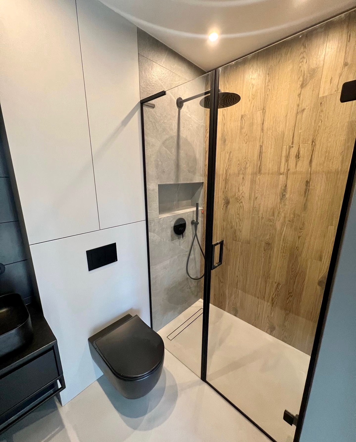 Nowoczesna łazienka z kabiną prysznicową typu walk-in, podwieszaną toaletą w kolorze czarnym i umywalką nablatową, wykończona płytkami imitującymi drewno i szarym kamieniem.