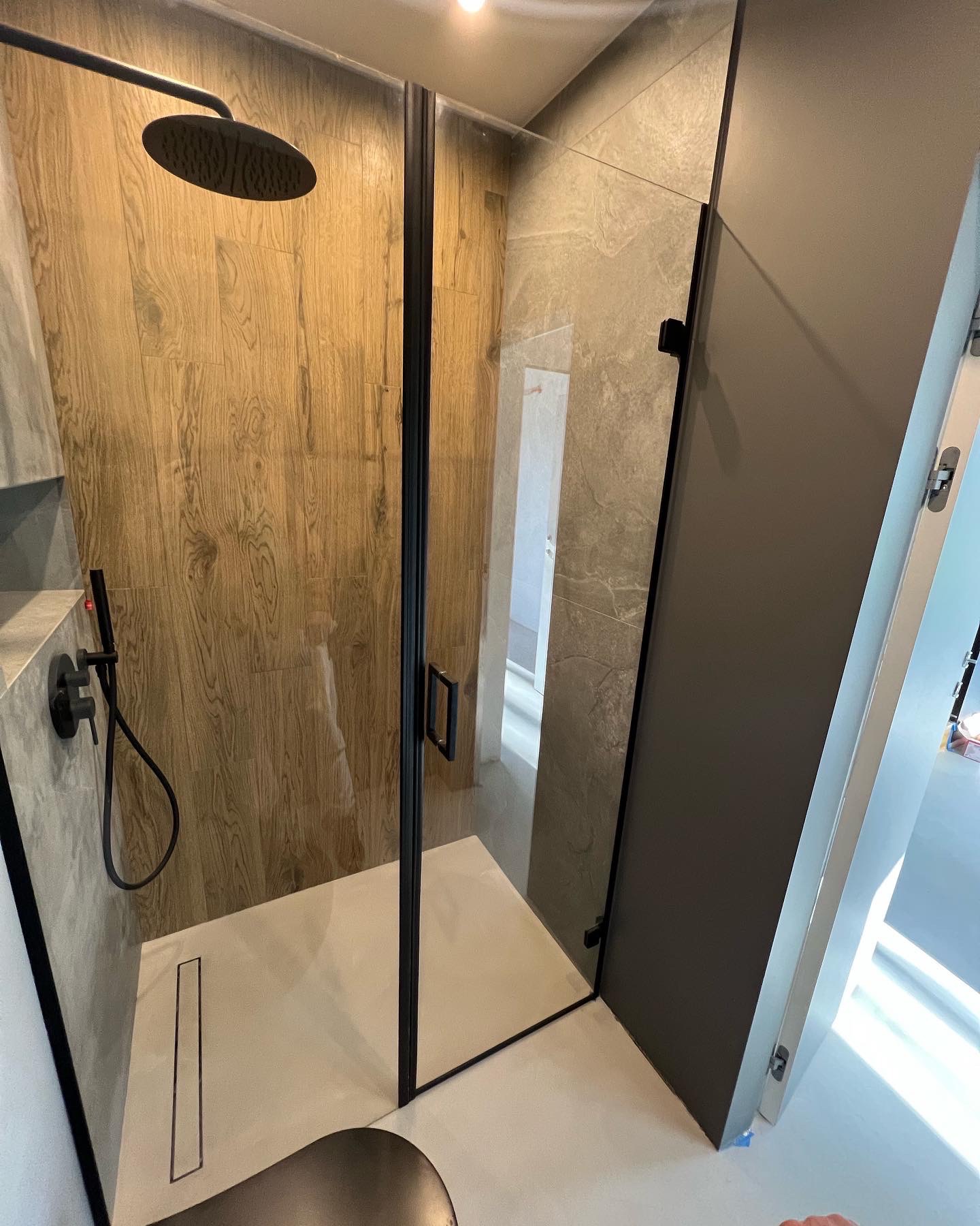 Nowoczesna kabina prysznicowa z czarnymi profilami, deszczownicą i odpływem liniowym, wykończona płytkami imitującymi drewno i kamień.