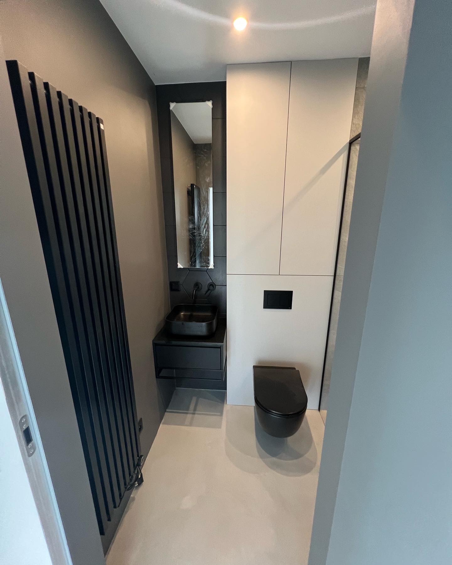 Nowoczesna łazienka w szaro-białej kolorystyce z czarnymi akcentami: grzejnik drabinkowy, umywalka nablatowa, wisząca toaleta. Umywalka z czarną armaturą i lustrem w czarnej ramie. Minimalistyczne...