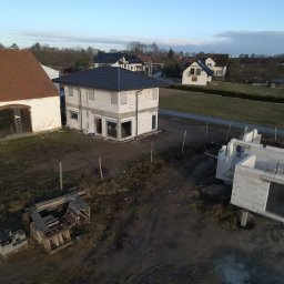 Urbzurek - Widok z góry na budowę domu z białych bloczków, z częściowo ukończoną konstrukcją i dachem, obok starszy budynek z ceglanym dachem, w tle zabudowa wiejska.