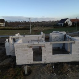 Urbzurek - Surowy stan domu w trakcie budowy, ściany z betonu komórkowego z widocznymi otworami okiennymi i drzwiowymi, na tle wiejskiej zabudowy i pochmurnego nieba.