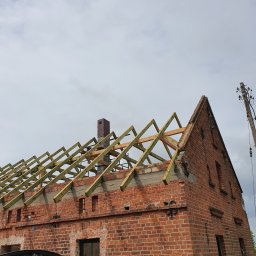 Urbzurek - Ceglany budynek w trakcie budowy dachu, widoczne drewniane krokwie, komin z cegły i fragment samochodu w lewym dolnym rogu.