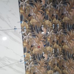 Urbzurek - Układanie płytek ściennych: białe imitujące marmur sąsiadują z płytkami w tropikalne liście i kwiaty, z użyciem klipsów poziomujących.