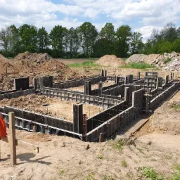 Zarys fundamentów domu jednorodzinnego z szalunkiem systemowym, widoczne zbrojenie, w tle hałdy ziemi i drzewa.