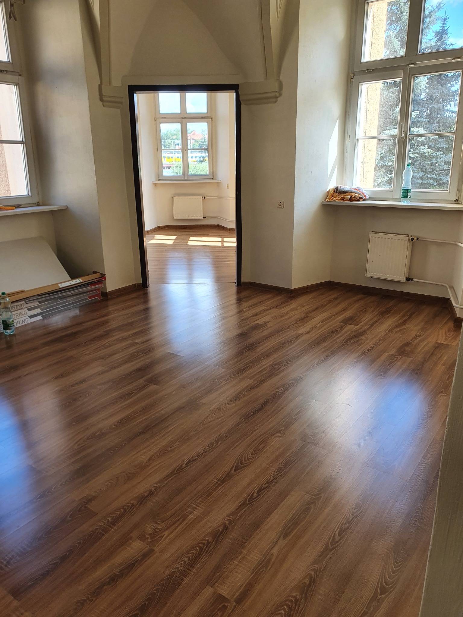 Ukończona podłoga z paneli drewnopodobnych w jasnym pomieszczeniu z wysokimi oknami, widoczne paczki paneli przy ścianie i butelka wody mineralnej.