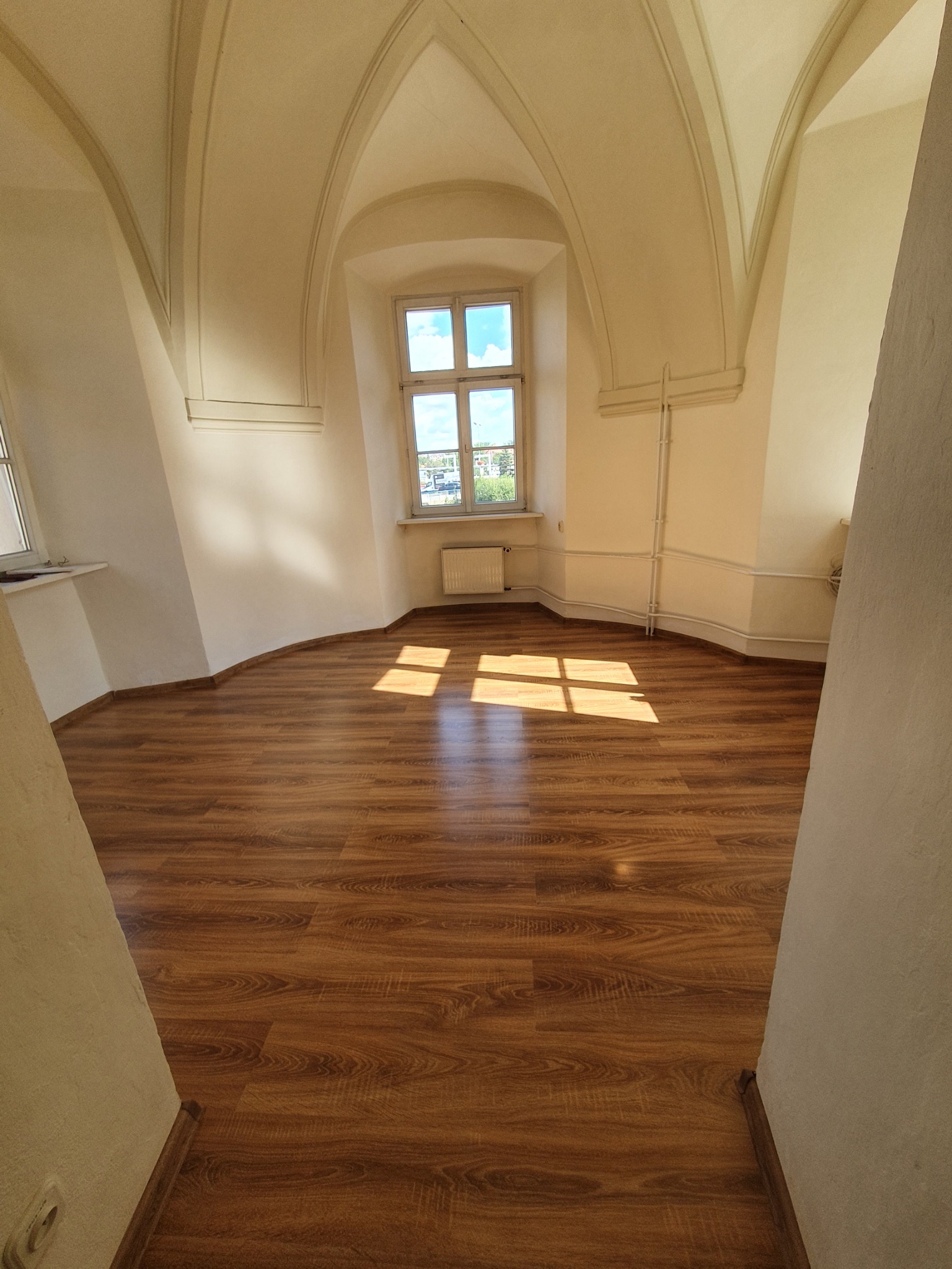 Puste pomieszczenie z łukowym sufitem i drewnianą podłogą w odcieniach brązu, oświetlone promieniami słońca wpadającymi przez okno.