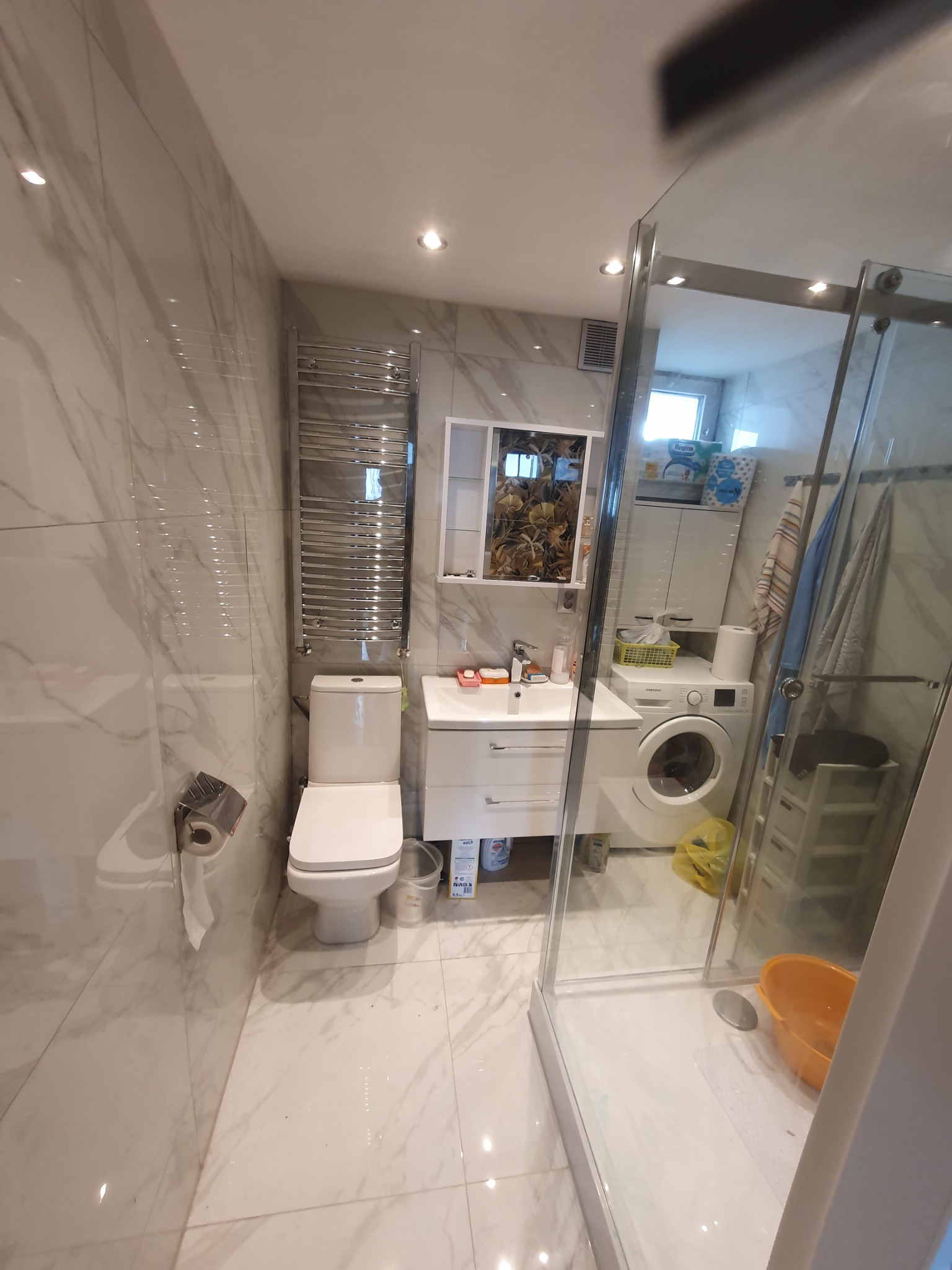 Jasna łazienka z marmurowymi płytkami, widoczny sedes, umywalka z szafką, grzejnik drabinkowy, kabina prysznicowa oraz pralka w tle.