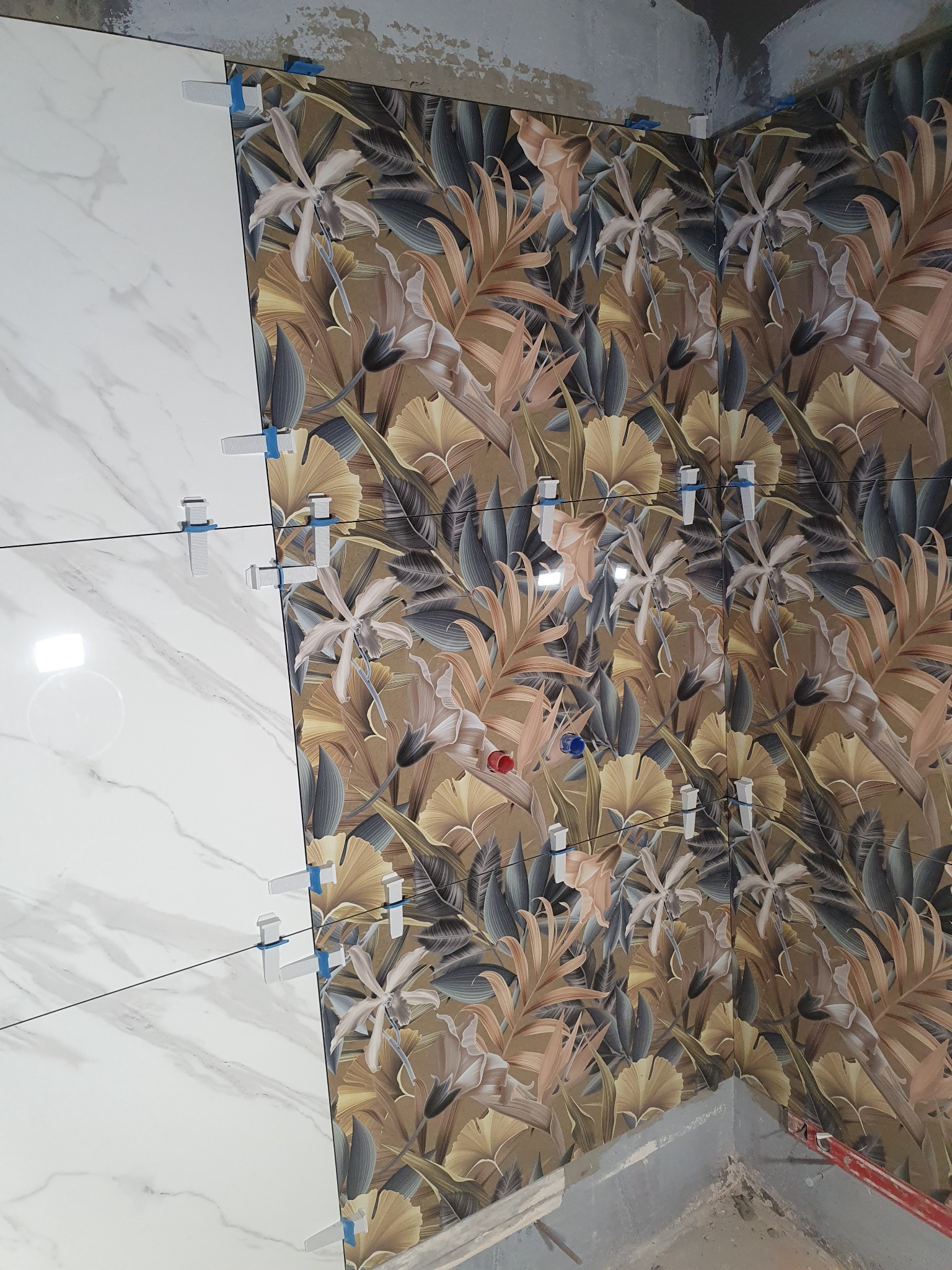 Układanie płytek ściennych: białe imitujące marmur sąsiadują z płytkami w tropikalne liście i kwiaty, z użyciem klipsów poziomujących.