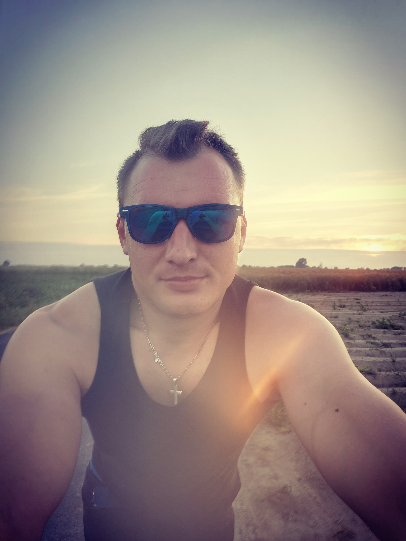 Mężczyzna w okularach przeciwsłonecznych i podkoszulku na tle zachodzącego słońca nad polem, selfie z dołu.