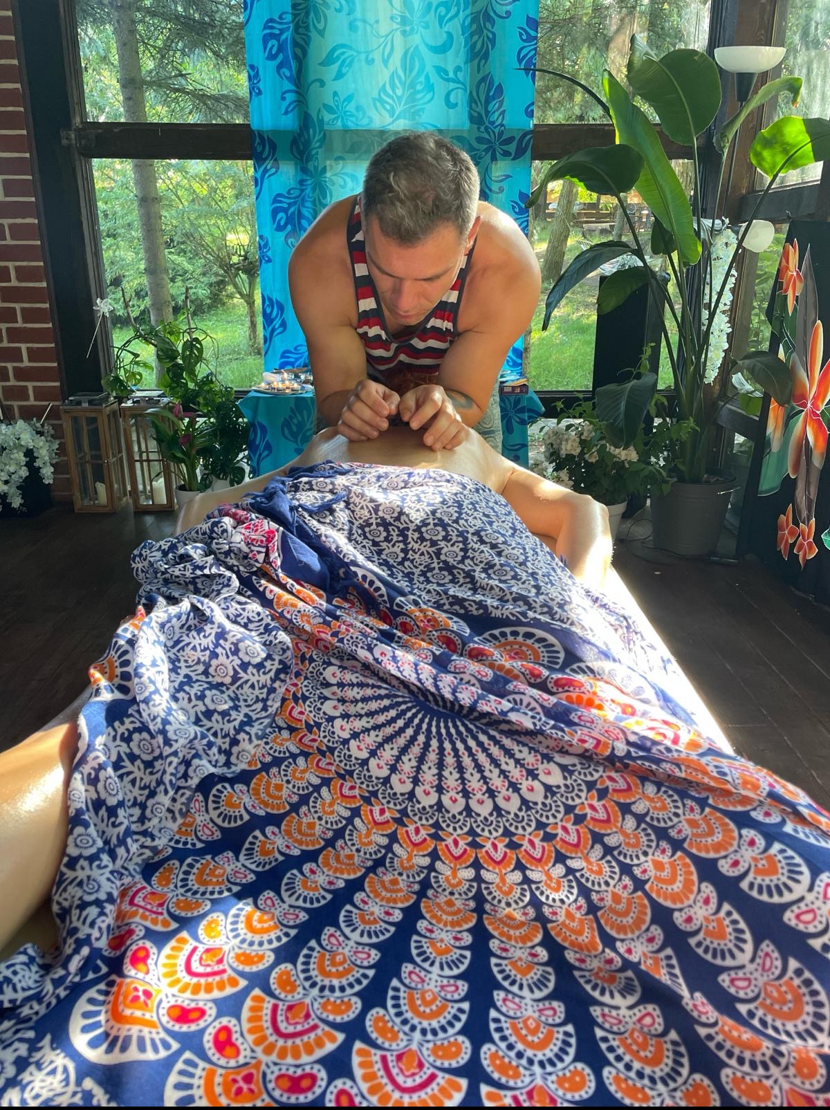 Mężczyzna w koszulce w paski wykonuje masaż pleców kobiecie leżącej na stole przykrytym kolorową tkaniną, w tle okno z zielenią i dekoracje roślinne.