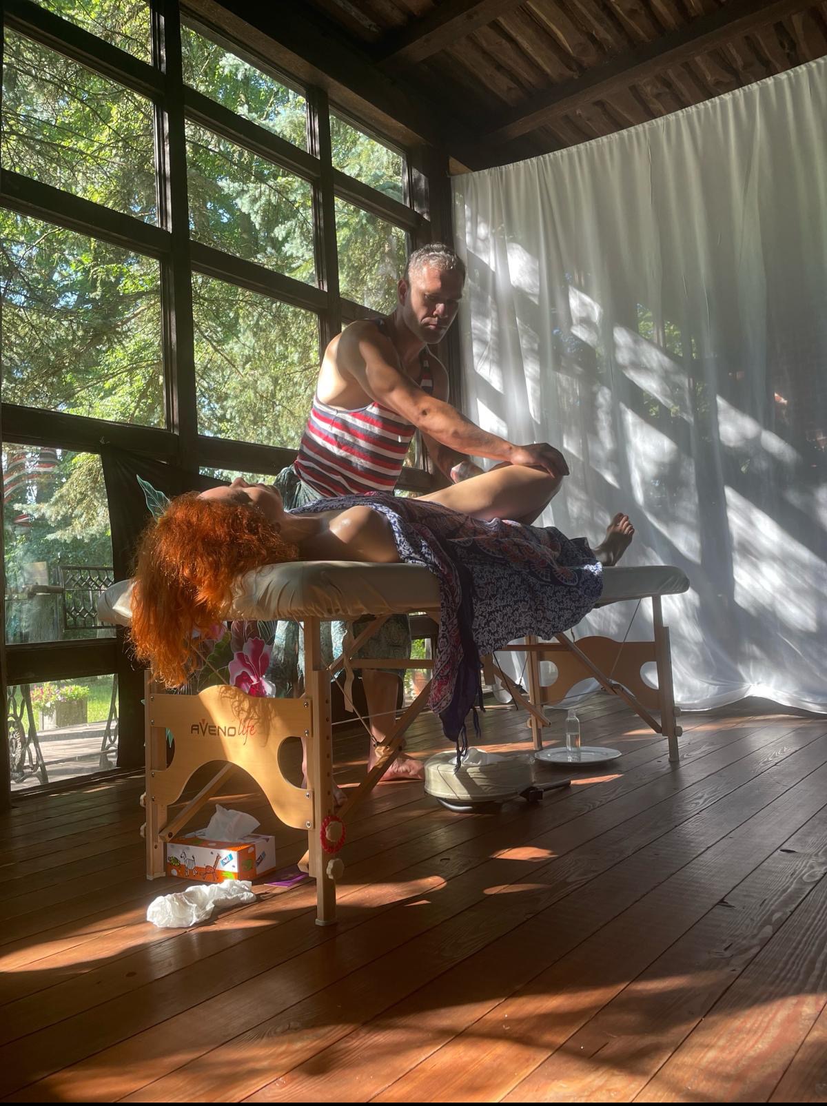 Mężczyzna w pasiastym podkoszulku wykonuje masaż nóg u kobiety leżącej na drewnianym łóżku do masażu w jasnym pomieszczeniu z dużymi oknami.