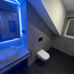 Nowoczesna łazienka z szarą ścianą, czarną podłogą i oświetleniem LED w kolorze niebieskim, widoczna umywalka nablatowa i toaleta podwieszana.